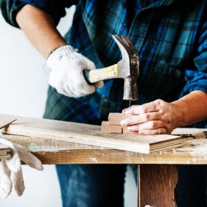 Image contains a man hammering a nail in wood.